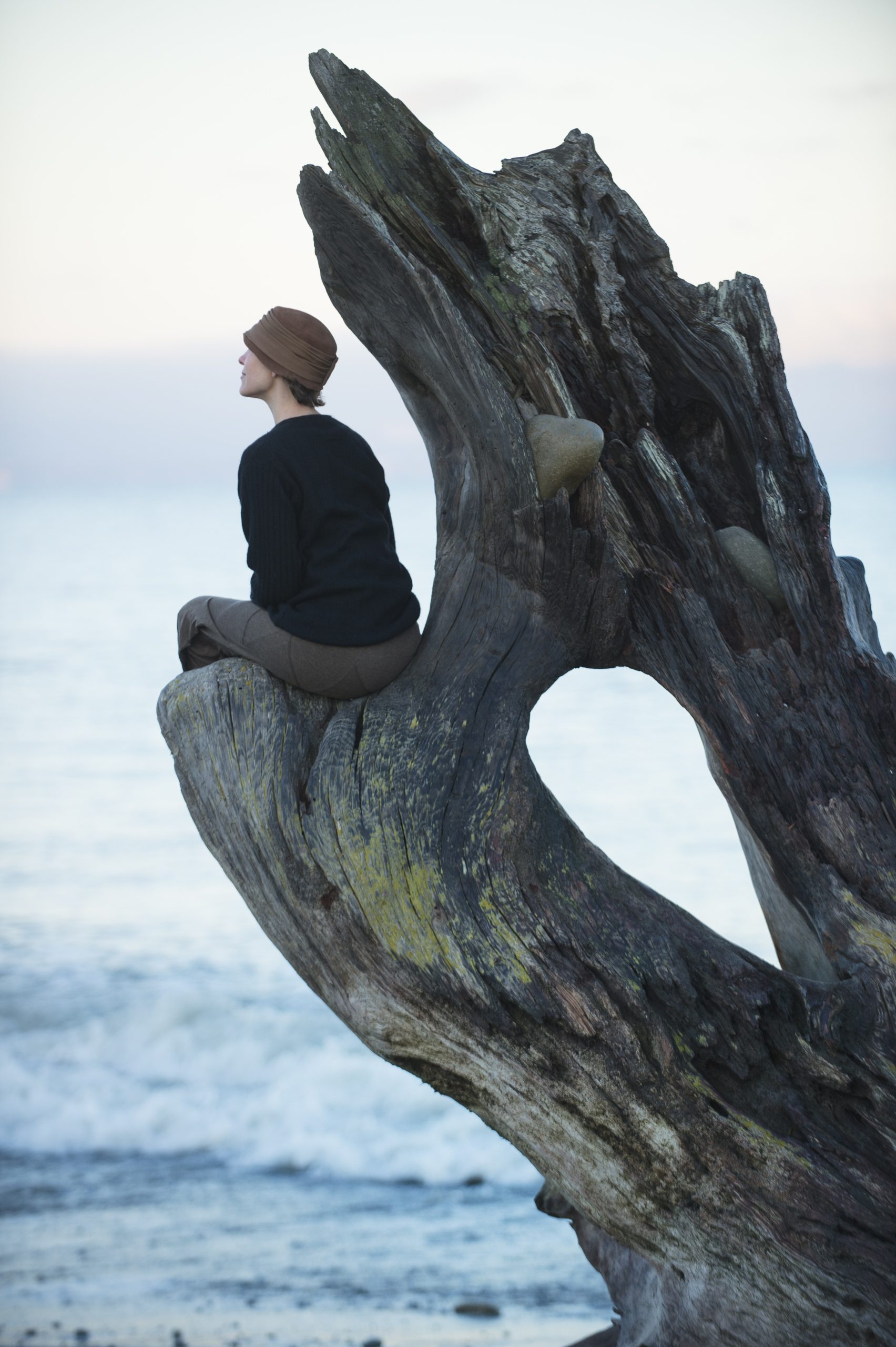 Woman sitting looking out from large driftwood tree trunk on beach